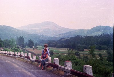 Two women seated on a roadside barrier overlooking a valley of lush, forested hills. View is dominated by a distant, hazy mou...
