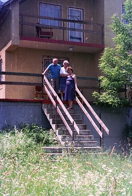 A candid, color photograph captures an elderly couple on the concrete stairs of a 1970s apartment block. Man in a short-sleev...