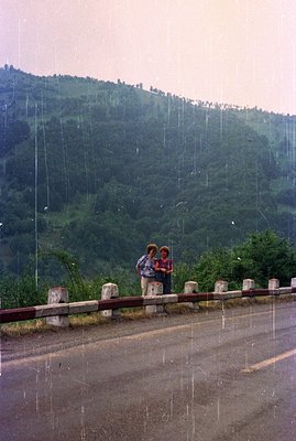 Two figures stand near a roadside barrier overlooking a heavily forested hillside under a rainfall. The scene suggests a moun...