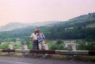 An older couple stands behind a road safety barrier, overlooking a forested valley with a river winding through it. Man wears...