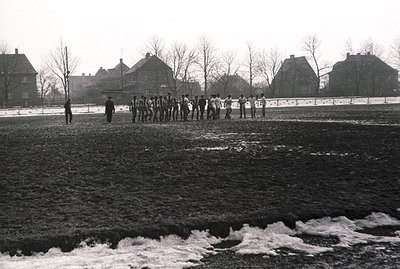 A group of young men, possibly a sports team, stand in a line on a muddy field with a row of brick buildings in the backgroun...