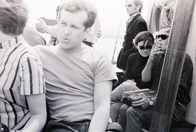 Three men aboard a sailboat, appearing relaxed and engaged in smoking. The central man wears a simple t-shirt, while others s...