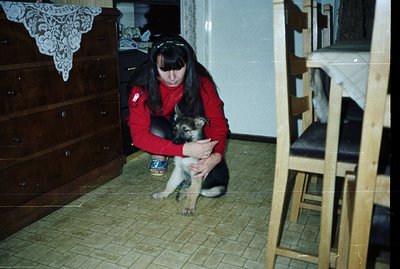 A young woman in a red sweater kneels, cradling a German Shepherd puppy indoors. Visible are a dresser with lace detailing, a...