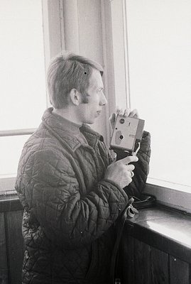 Man in a quilted jacket operates a vintage rangefinder camera, positioned near a window. Likely a candid shot, capturing a mo...