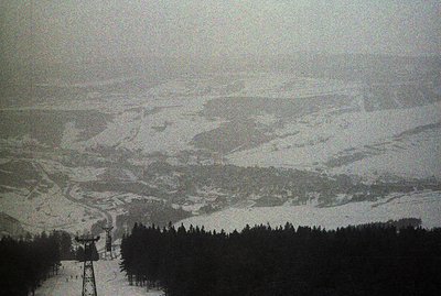 An aerial view captures a snow-covered alpine landscape featuring a ski lift tower and groomed slopes. Dense evergreen forest...