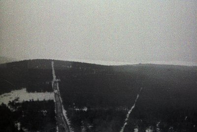 A high-angle view captures a ski lift ascending a forested, snow-covered slope. The stark monochrome highlights the infrastru...