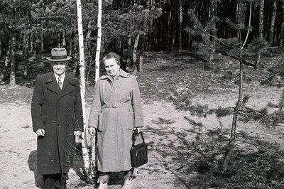A formally dressed couple stands beside a birch tree within a dense pine forest. The man wears a dark overcoat and hat; the w...