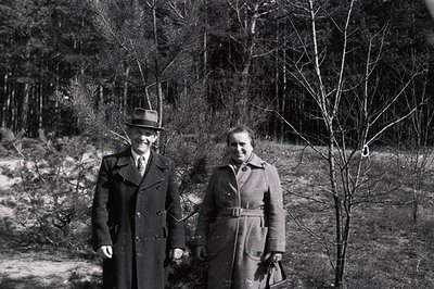A formally-dressed man and woman stand outdoors, likely in a park or forest setting. Both wear long overcoats and hats, sugge...