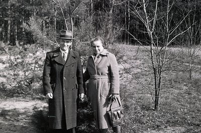 A formally-dressed couple stands outdoors in a sparse, wooded area. The man wears a double-breasted coat, fedora, and tie; th...