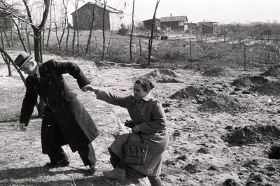 A man in a long coat and hat assists a woman in a coat and gloves, who appears to be struggling on uneven, muddy ground. Like...