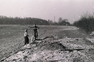 A young man and a child stand near a freshly-dug landscape, likely a construction site or excavation. Earth mounds are promin...
