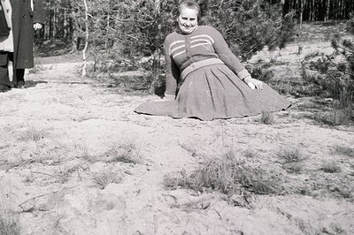 A woman in a textured sweater and full skirt poses in a sandy, grassy landscape, partially obscured by pine trees. A person i...