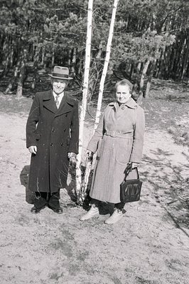 A formally-dressed couple stands near birch trees in a wooded area. The man wears a long overcoat and fedora; the woman, a be...