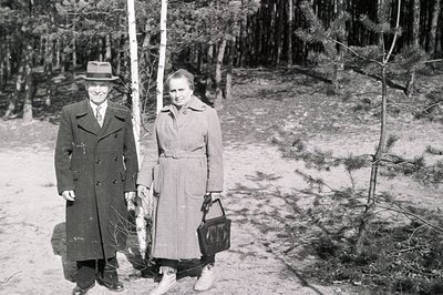 A formally dressed man and woman pose near birch trees in a wooded area. The man wears a double-breasted coat, fedora, and ti...