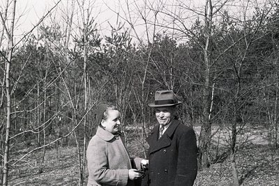 A formally-dressed couple stands amongst bare trees in a wooded area, likely late fall or winter. The woman wears a long coat...