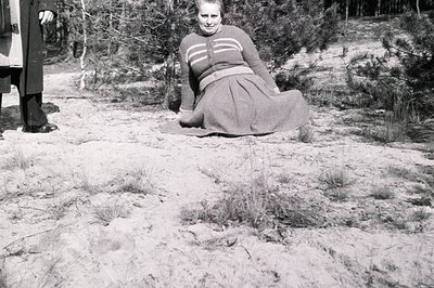A woman in a full, pleated skirt and sweater sits on a sandy area, partially obscured by pine trees. A man in a dark coat and...