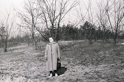 A woman in a long coat stands on a grassy slope, holding a dark bag. Bare trees populate the background, suggesting late wint...
