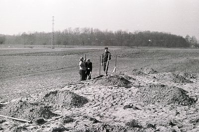 Dusty, monochromatic image depicts a construction site with three figures amidst disturbed earth and piled soil. A man stands...