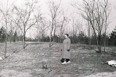 A woman in a long coat stands on a grassy slope, framed by bare trees. Likely a mid-century outdoor portrait, showcasing a st...