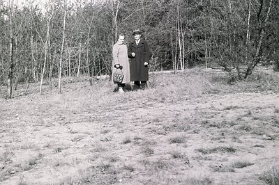 A man and woman stand on a grassy, gently sloping hillside under bare trees. Both are dressed in 1930s-era clothing: the woma...