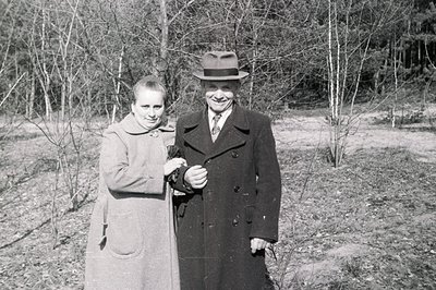 A formally-dressed couple poses outdoors amidst leafless trees, likely in a park setting. The woman wears a long coat with a ...