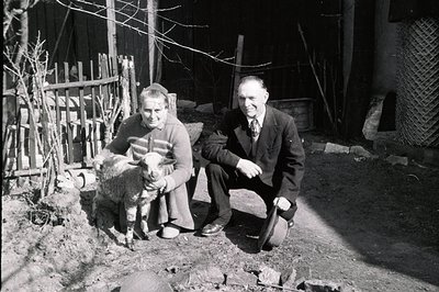 A middle-aged couple poses with a young lamb in a rural courtyard. The woman wears a sweater and long skirt; the man, a suit ...
