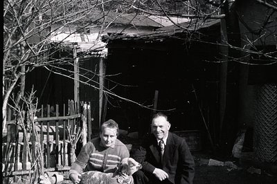 A man and woman, formally dressed, sit with a young lamb in front of a weathered, simple structure, possibly a shed or barn. ...