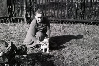 A seated woman cradles a kitten outdoors near a woodpile. She wears a dark, knit sweater and skirt. The backdrop features a w...