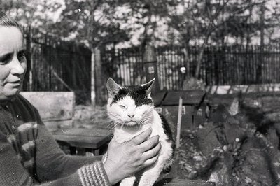 A woman gently holds a black and white cat; both appear to be looking directly at the camera. The scene suggests an outdoor s...