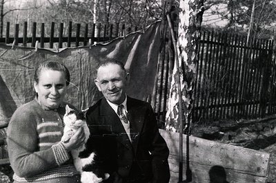 A couple poses in a backyard setting, woman holding a cat. Man wears a dark suit and tie. Rustic wooden fence & tarp backdrop...