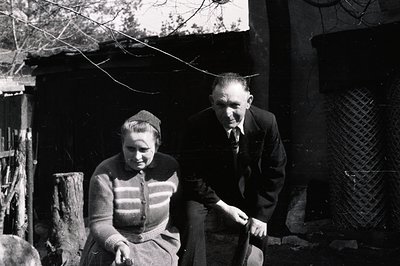 A man and woman sit outside a rustic, whitewashed building with a timbered roof. The man wears a suit and tie; the woman a ca...