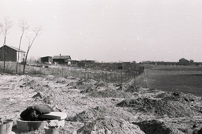 A laborer rests on a makeshift platform amid construction debris and excavated earth. A barbed-wire fence separates the const...
