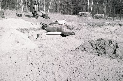 Black and white image documenting a suspected excavation site. A body lies on a wooden board within a newly-dug trench. Sever...