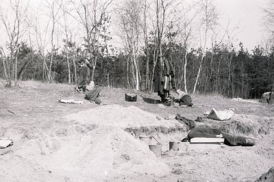 Archaeological excavation in progress. Visible are several workers carefully uncovering skeletal remains within a sandy, slop...