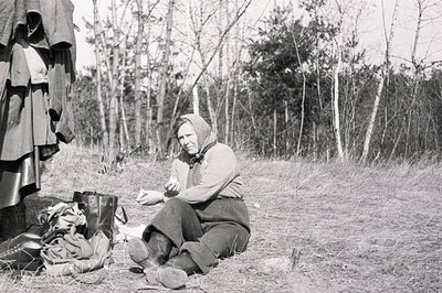 A woman in traditional attire sits outdoors amongst scattered belongings and a hanging garment. She wears a headscarf, dark s...