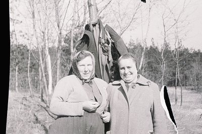 Two women in winter coats & headscarves pose outdoors, likely in a rural setting. Appears to be a candid portrait, possibly a...