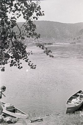 A lakeside scene: a woman stands near a boat on a pebbled shore, with hills rising in the background. Two rowboats sit on the...