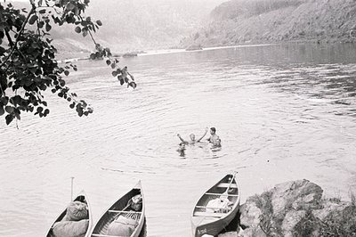 Three wooden canoes are beached near a shoreline with two figures swimming in a wide, placid river framed by steep, forested ...