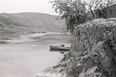 A lone canoe sits on a calm river, with a person visible inside. Rocky shoreline and a tree overhang the water's edge. Rollin...