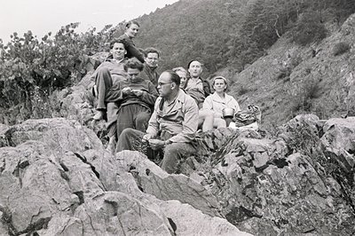 A group of six people pose amidst a rocky, overgrown hillside, likely during a hike or outing. Men wear work shirts & some sp...