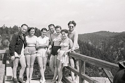 A group of six people pose on a wooden balcony overlooking a forested hillside. Mid-century swimwear and casual wear suggest ...