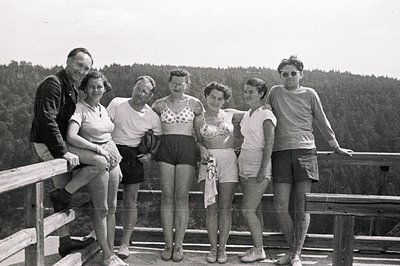 Group portrait, six individuals posing on a balcony overlooking a dense forest. Appears to be a seaside resort or vacation sp...
