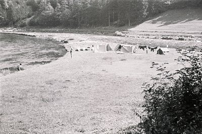 A riverside campsite features several canvas tents arranged along a grassy bank. A lone figure stands near the shoreline. Den...
