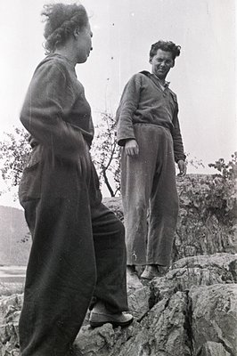 Two figures are captured on a rocky, coastal ledge. A woman in wide-legged trousers and a loose-fitting top stands observing ...