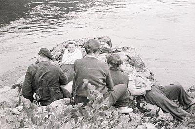 Four people, partially obscured, relax on a rocky outcrop beside a body of water. Three wear jackets and trousers; one wears ...