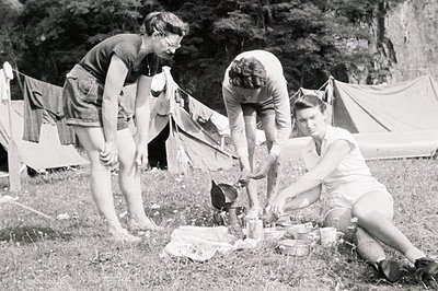 Three young people prepare food outdoors near a tent, seemingly on a camping trip. One is barefoot, another is tending to a s...
