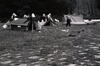 A grassy field houses several military-style tents, with laundry lines strung between them. A soldier stands near a tent, see...