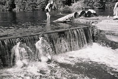 Two women in swimwear pose near a small waterfall, enjoying a riverside recreation area. A young man is jumping into the wate...