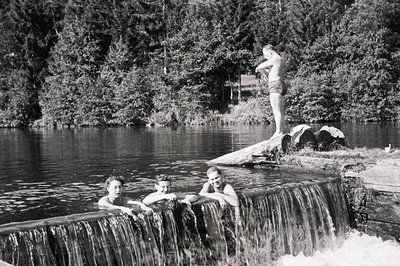 Three young men enjoying a summer swim at a small dam/waterfall. Two are partially submerged, seated atop the concrete struct...
