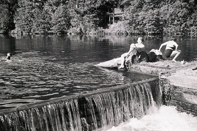 Three young people relax on a stone ledge beside a small waterfall and reservoir. Two are reclining, one appears to be swimmi...